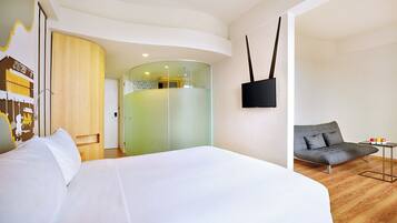 A modern hotel room with a large bed, a sofa, a flat-screen TV, and a glass partition wall.