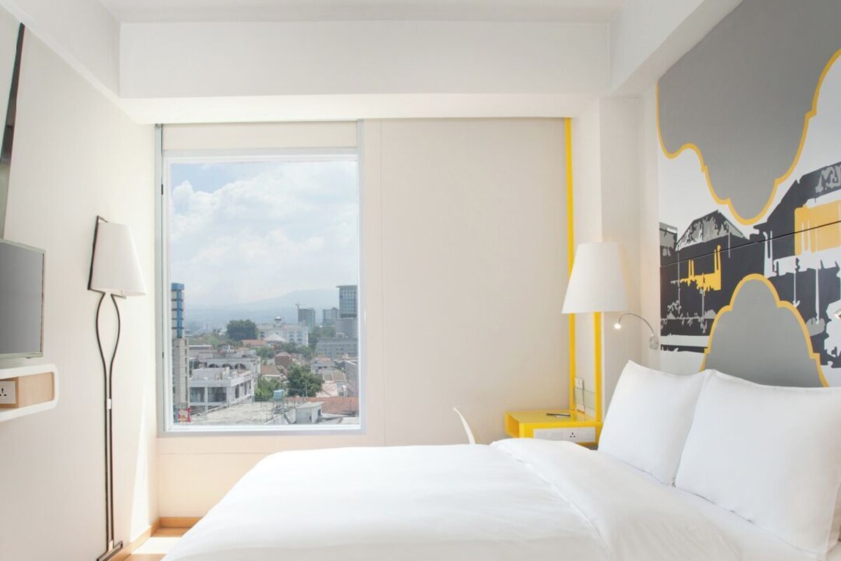 Une chambre d’hôtel avec un grand lit, une vue sur la ville, un bureau avec une lampe et un mur orné d’un motif graphique.