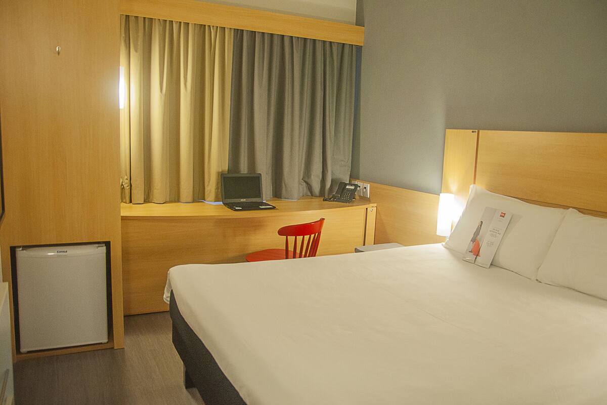 Habitación de hotel con cama, escritorio con computadora portátil, silla y refrigerador pequeño.