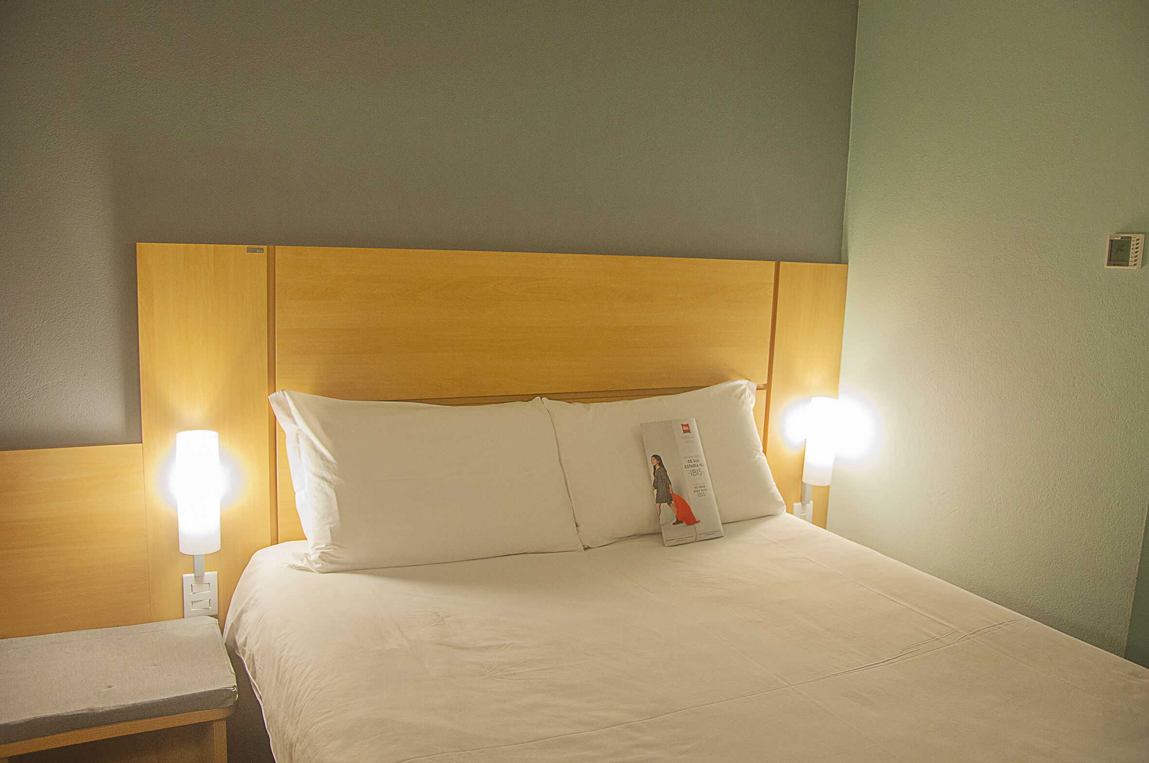 A hotel room with a wooden headboard, two bedside lamps, a white bed with pillows, and a bedside table.