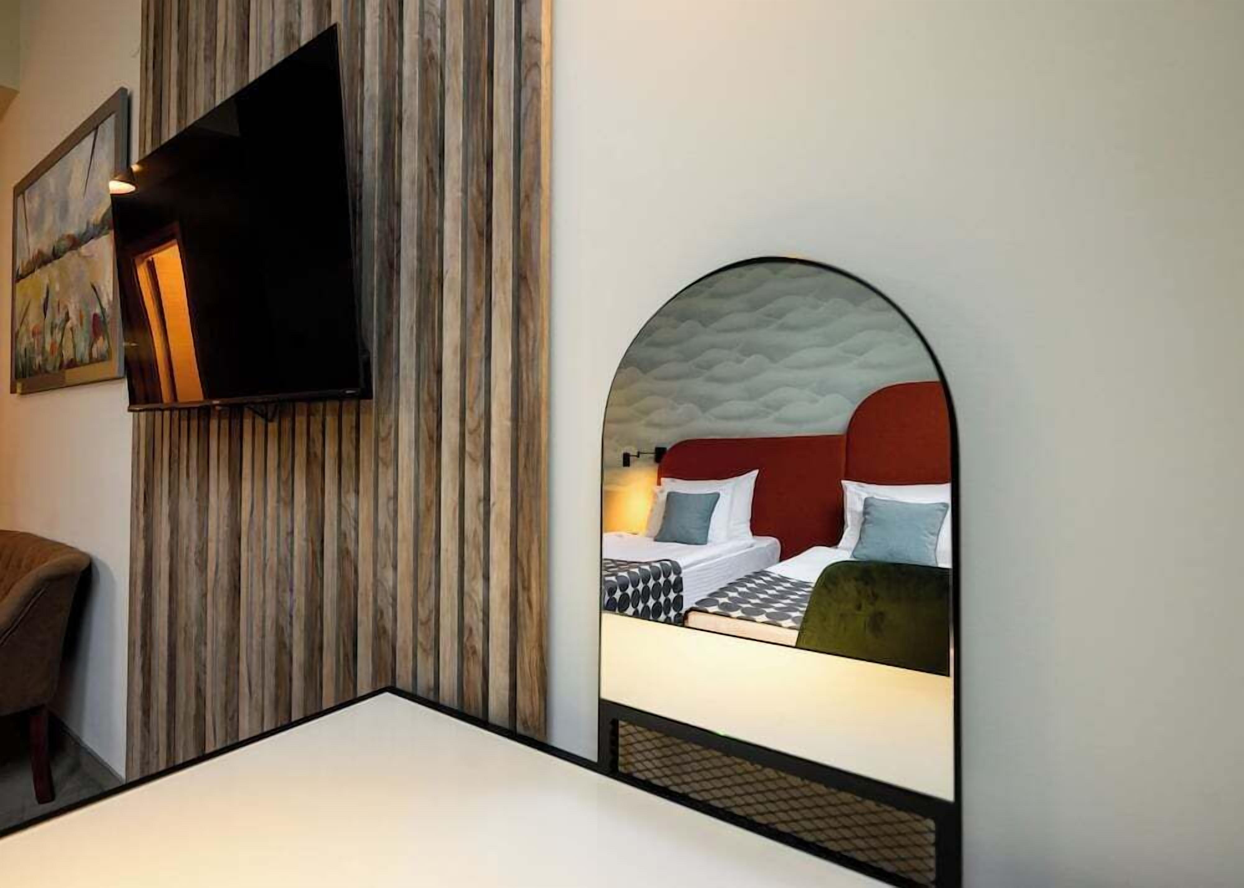A hotel room with a television mounted on the wall, a bed with a red headboard, and a large mirror reflecting the room.