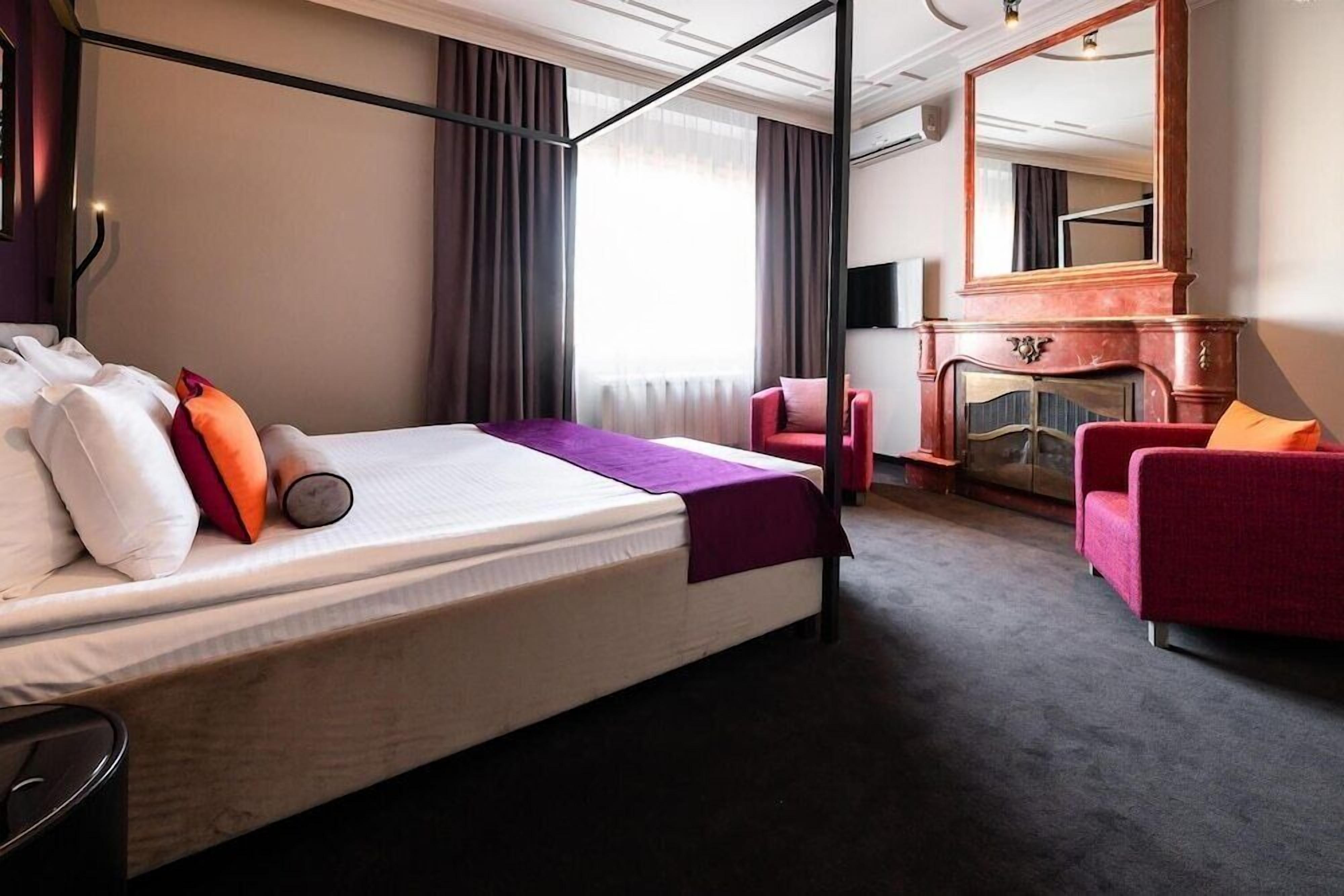 Una camera d'albergo con un letto, una poltrona rossa, un caminetto e un grande specchio.