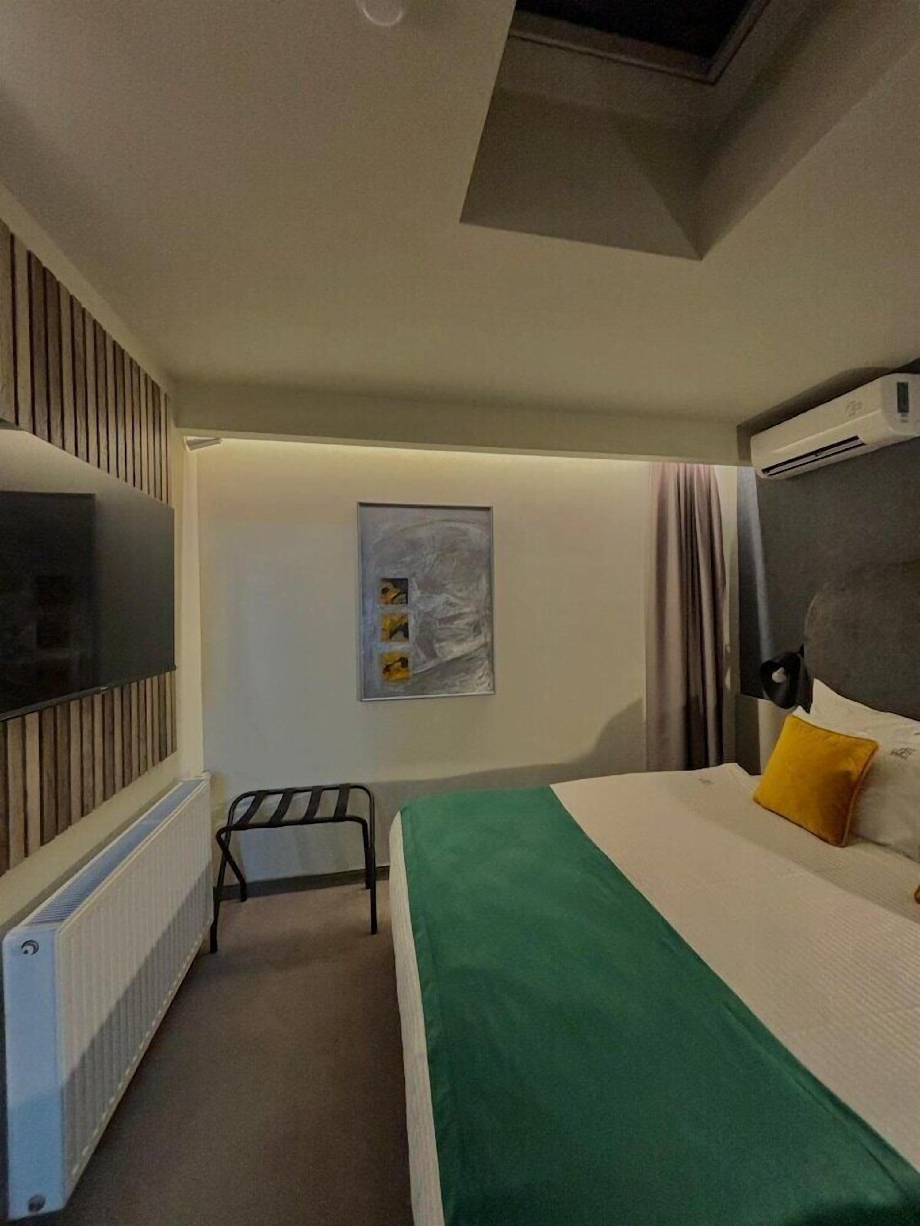 A hotel room with a bed, a radiator, a television, a picture on the wall, and a window with curtains.