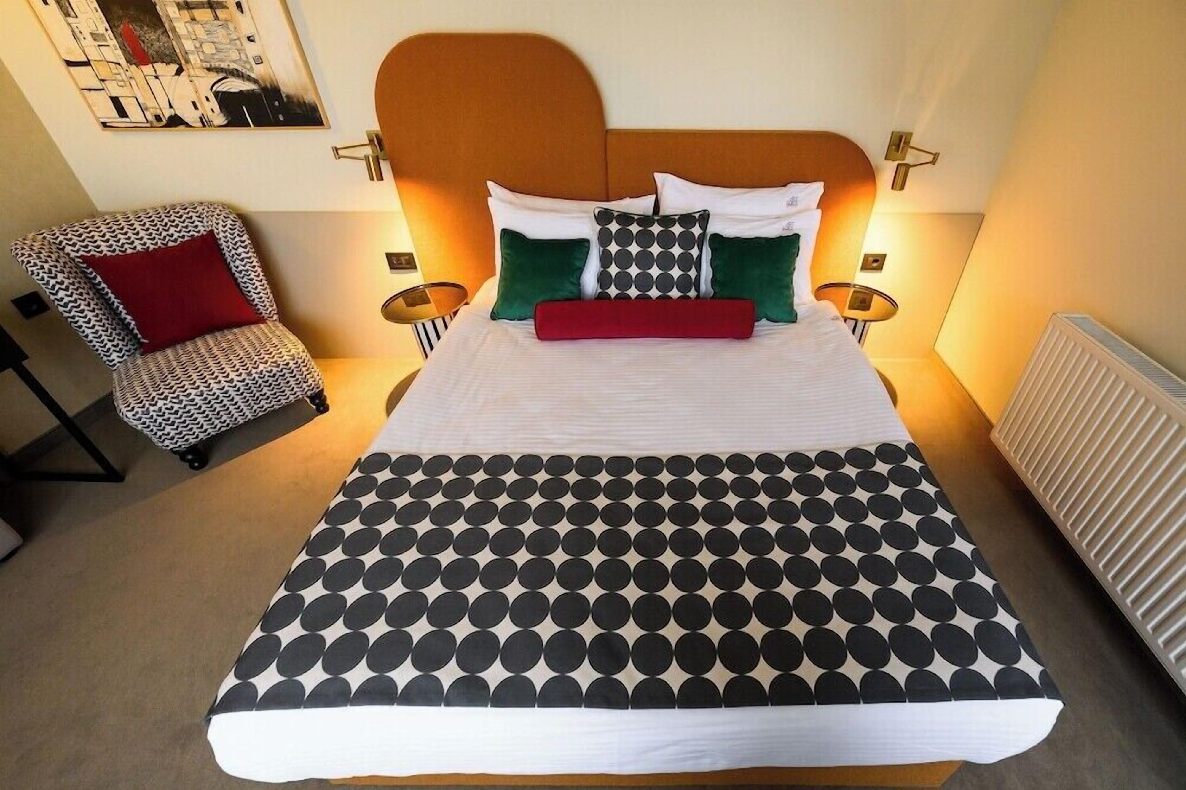 Uma cama com colcha estampada em padrão geométrico, uma cadeira com almofada estampada e um luminário fixado na parede.