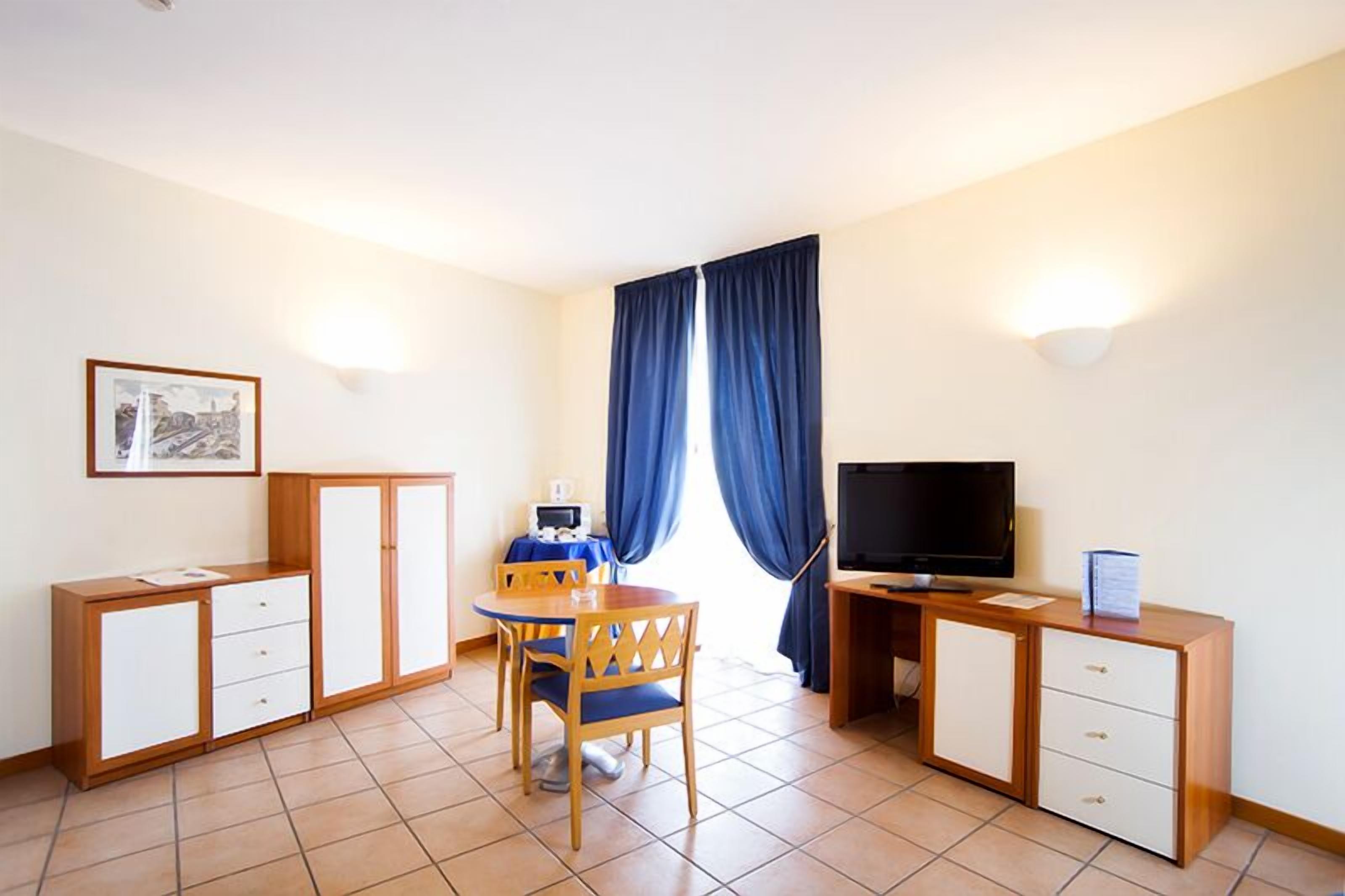 A small, clean room with a bed, a desk, a chair, a television, and a window with curtains.