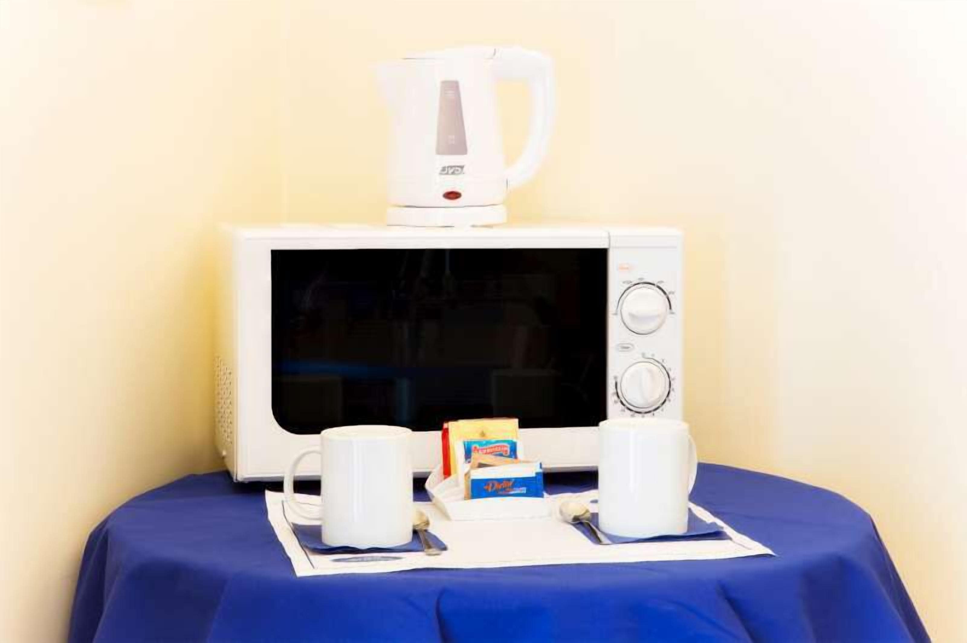 A microwave with a coffee pot on top, two white cups, and a box of tea on a table with a blue tablecloth.