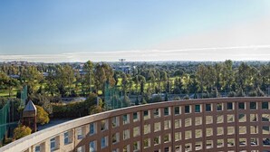 Une vue depuis un balcon donnant sur un paysage urbain, avec un ciel dégagé, comprenant un mélange de bâtiments résidentiels et commerciaux, ainsi qu’un parc avec des arbres et des allées.