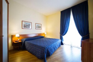 A hotel room with a large bed, two framed pictures on the wall, a nightstand with a lamp, and a window with curtains.
