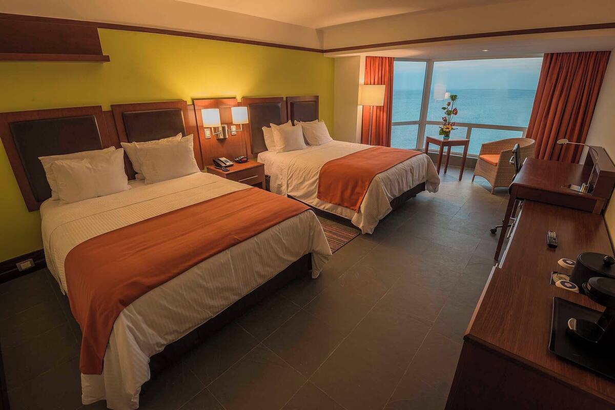 Habitación de hotel con dos camas, un escritorio, una silla y vista al mar.