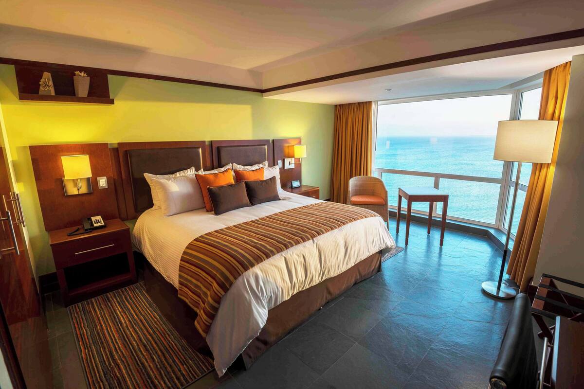 Habitación de hotel con una cama grande, una mesita de noche con lámpara, una silla y vista al océano.