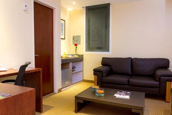 A compact living space with a dark brown leather sofa, a wooden desk with a chair, a small coffee table with decorative items, and a kitchenette area.