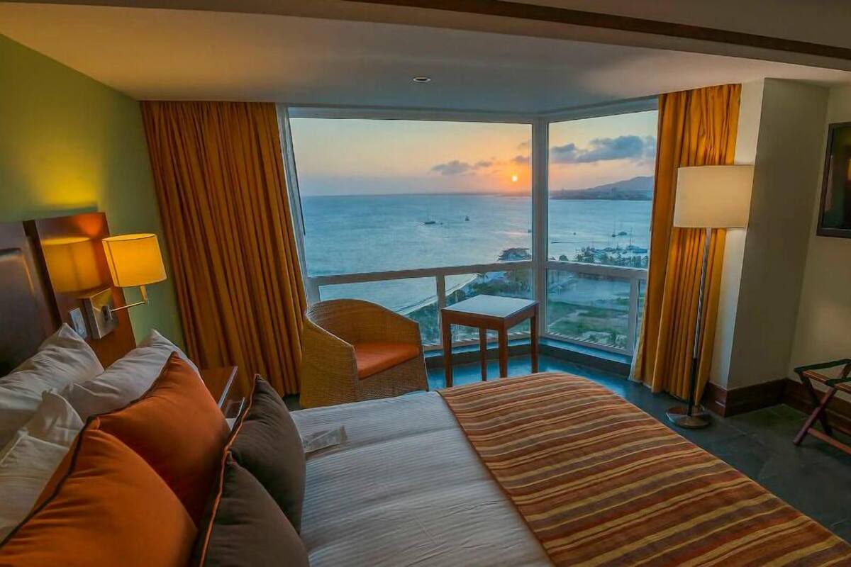 Habitación de hotel con una cama grande, una silla, una mesita, un televisor y un balcón con vistas al mar y al atardecer.