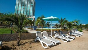 Un hotel frente a la playa con tumbonas y sombrillas, un edificio alto al fondo y palmeras.