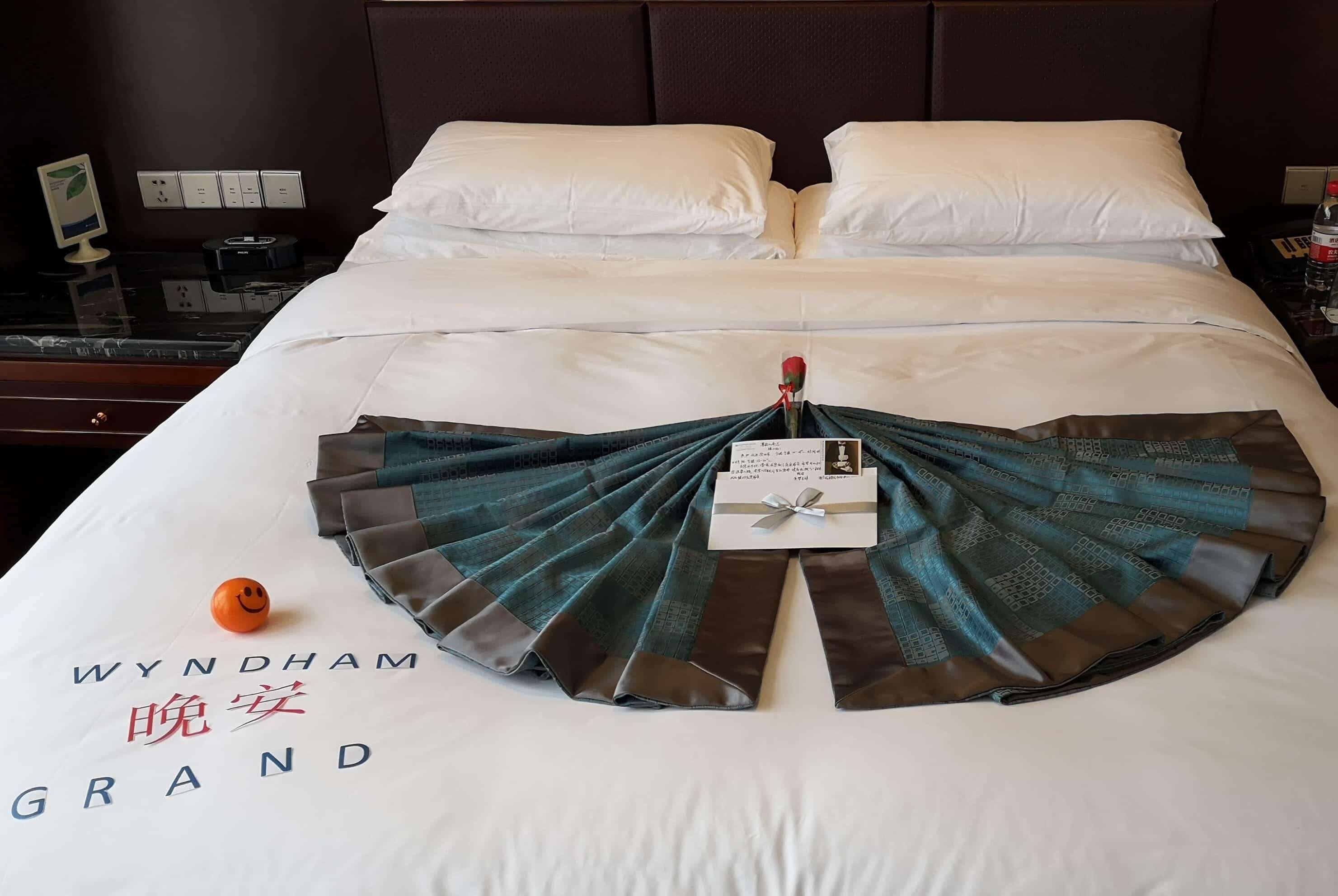 Un lit bien fait, agrémenté d’un éventail décoratif et d’une carte, d’un petit objet décoratif représentant un visage souriant, et sur lequel on peut lire le texte « WYNDHAM GRAND » brodé sur la couverture.