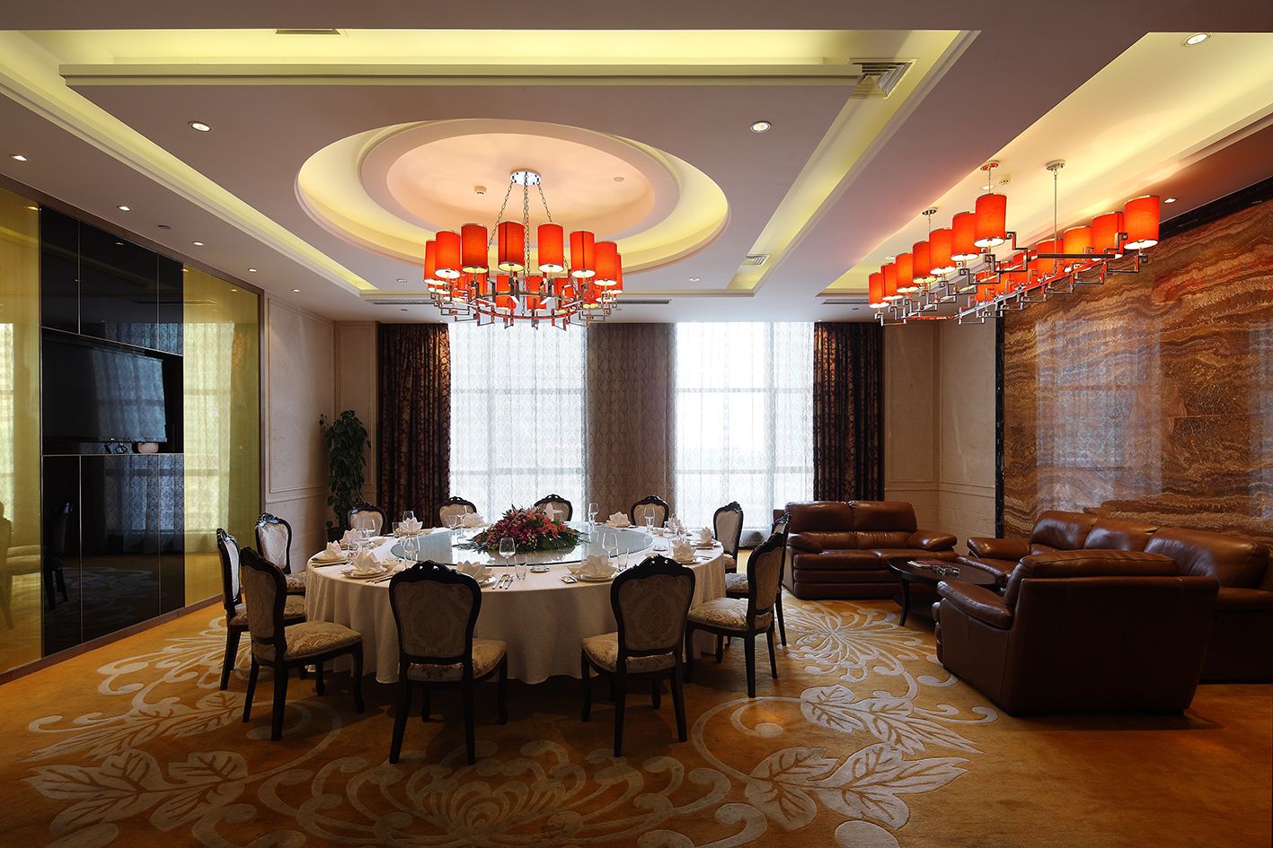 A formal dining room with a round table set for six, surrounded by chairs. There is a large window with curtains, a chandelier, and a leather sofa in the background.