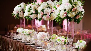 Ein langer Festtisch mit hohen floralen Dekorationen in klaren Glasvasen, gedeckt mit feinem Porzellan, Besteck und Weingläsern.