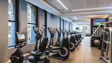 Un gimnasio con varias máquinas de ejercicio, amplias ventanas y una pantalla digital que muestra información sobre rutinas.