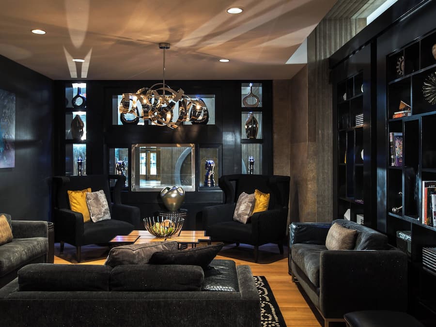 A modern living room with dark sofas, a coffee table, and a chandelier. There are bookshelves with decorative items and a large window.