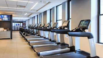 A row of treadmills in a gym with large windows and a TV screen in the background.
