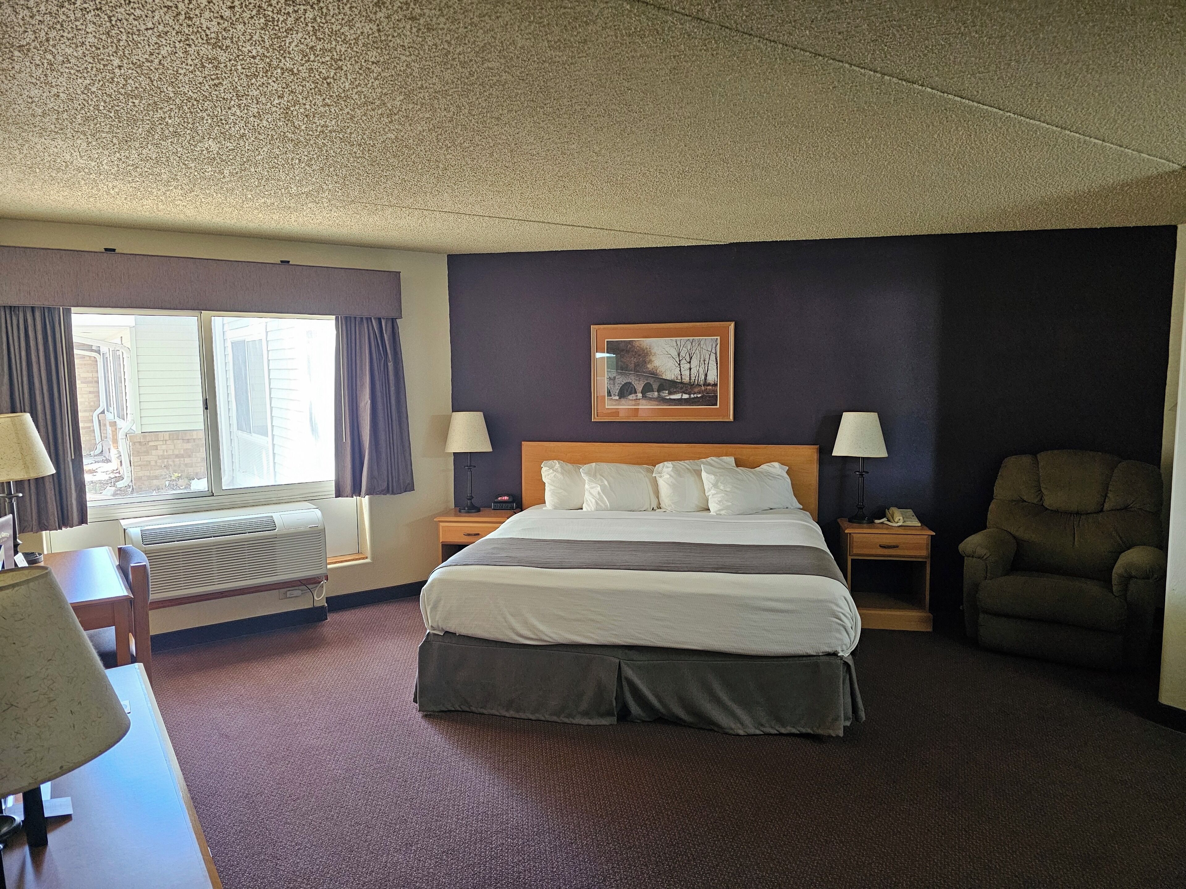 A hotel room with a large bed, two bedside tables with lamps, a chair, a window with curtains, and a framed picture on the wall.