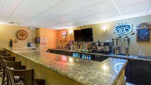 Uma área de bar com bancada de granito, banquetas, uma televisão e diversos frascos de bebidas alcoólicas.