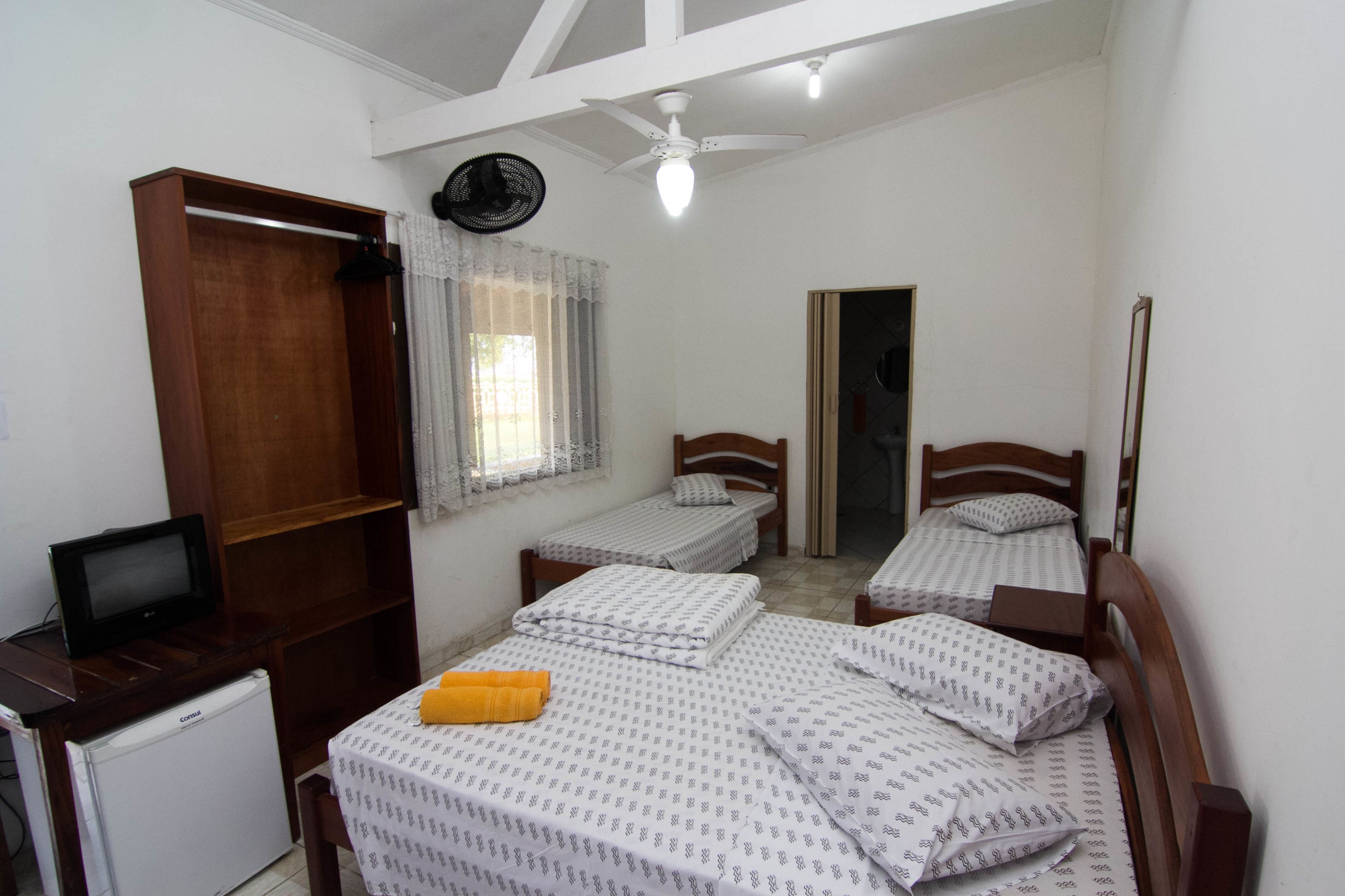 A small, well-lit bedroom with two single beds, a wooden wardrobe, a small desk with a television, and a ceiling fan.