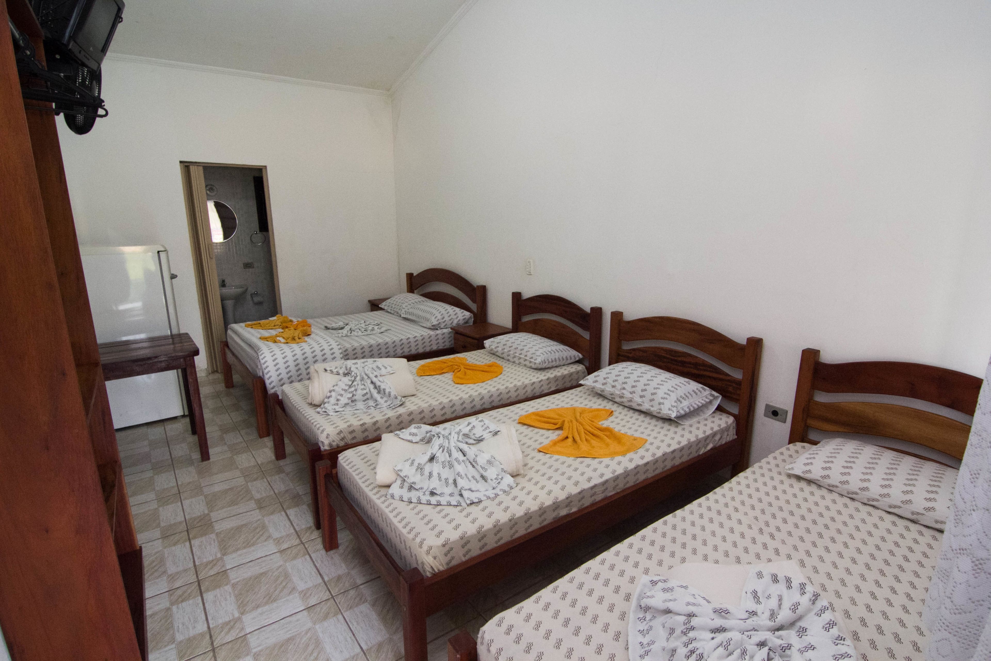 A dormitory room with four single beds, each with a wooden headboard and patterned bedding. A small table and a chair are visible in the corner.