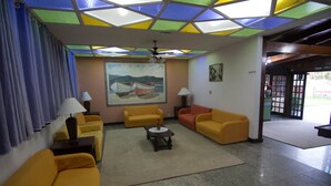 Ein Zimmer mit gelben Sofas, einem Couchtisch, einem Gemälde von Booten und einer Decke mit farbigen Glasfeldern.