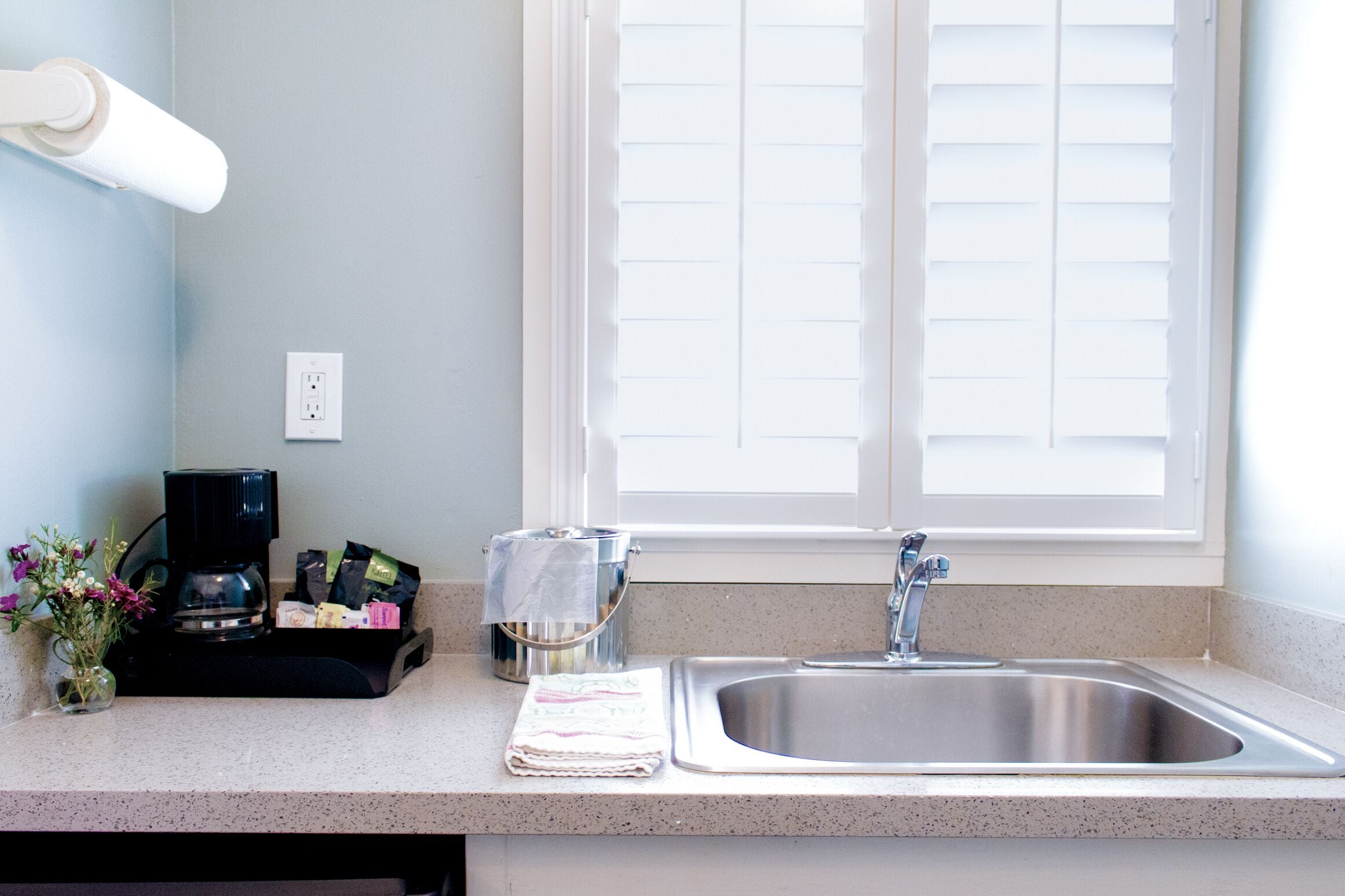 A kitchen with a stainless steel sink, a coffee maker, and a window with blinds.