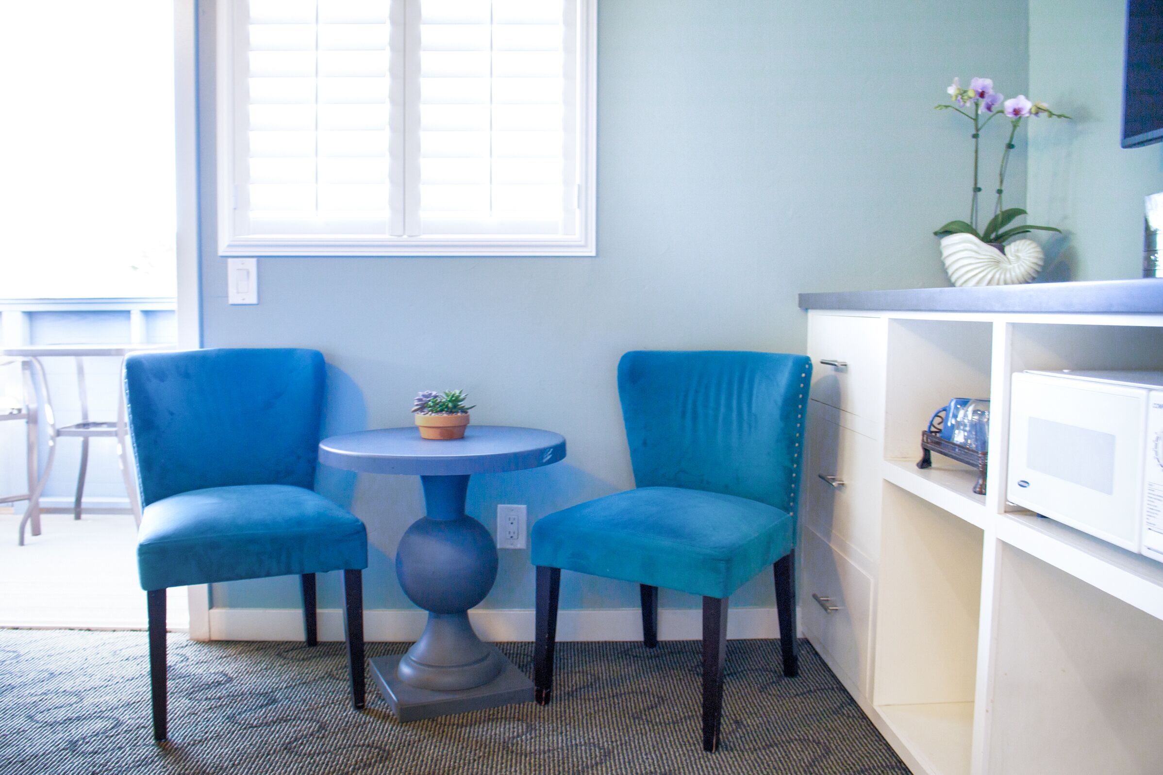 A room with two blue chairs, a small round table with a potted plant, a white cabinet with a microwave, and a window with blinds.