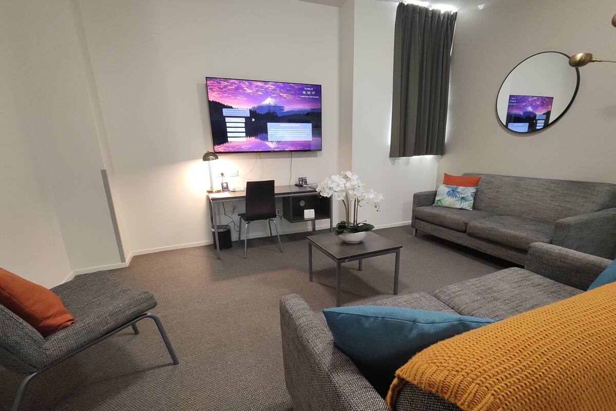 A modern living room with a grey sofa, a small table with a plant, a desk with a computer, and a TV displaying a scenic image.