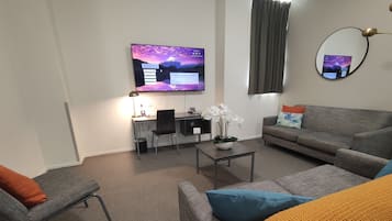 Una sala de estar moderna con un sofá gris, una mesita con una planta, un escritorio con una computadora y un televisor mostrando una imagen de paisaje.