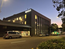 Un edificio con un letrero que dice "QUEST" en la parte superior, una fachada con un cartel que indica "Kathmandu", y varios autos estacionados en la calle.