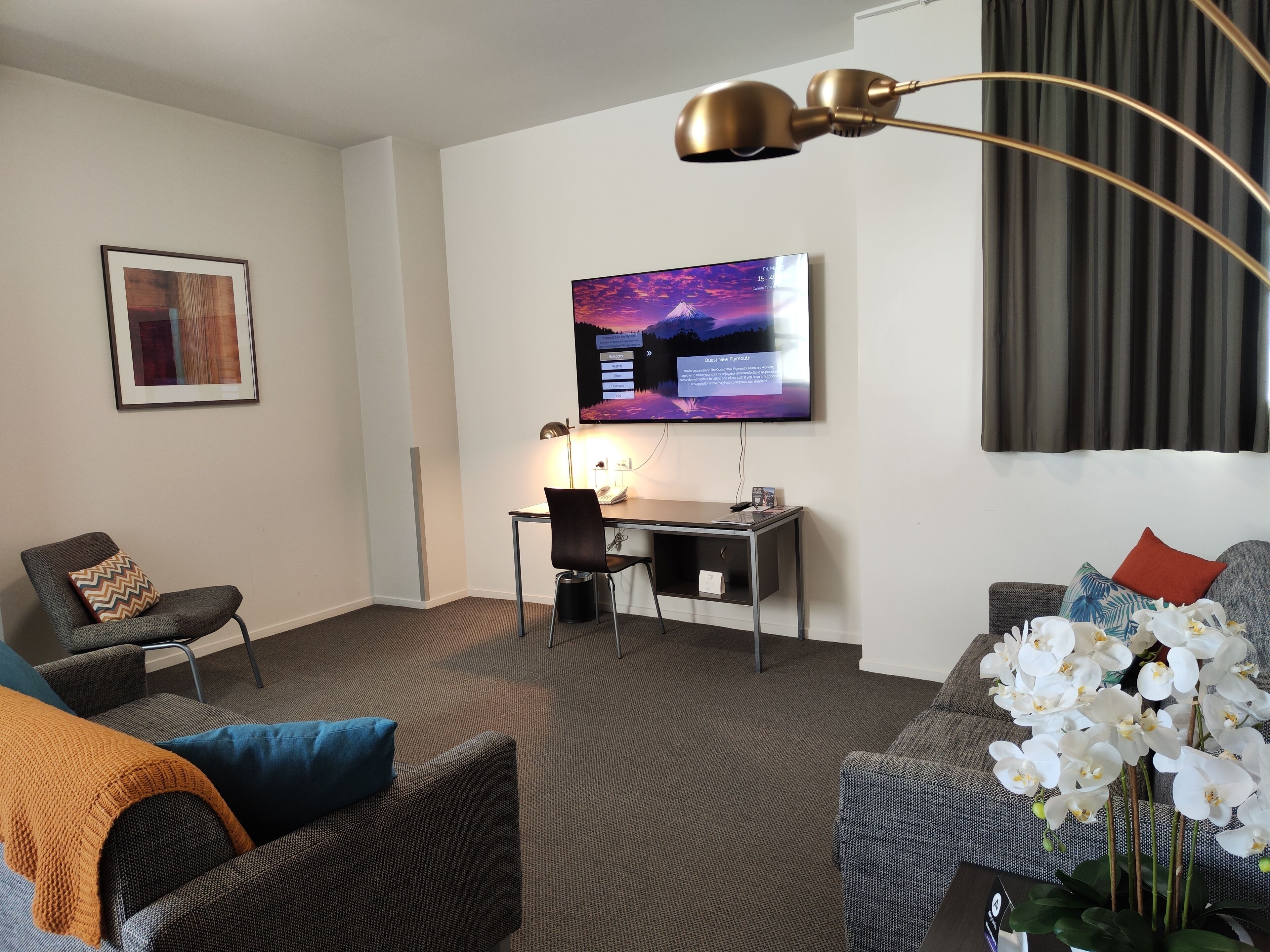 A modern hotel room with a flat-screen TV mounted on the wall, a desk with a chair, a sofa with a decorative pillow, and a large window with curtains.