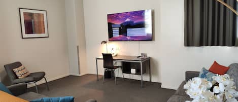 Habitación de hotel moderna con televisor de pantalla plana en la pared, un escritorio con silla, un sofá con almohadón decorativo y un ventanal con cortinas.
