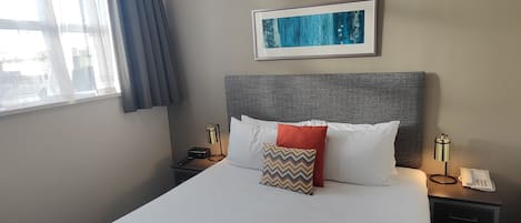 A neatly made bed with a grey headboard, white and red pillows, and a patterned bedspread. There is a framed picture on the wall above the bed, and a bedside table with a lamp and a phone.