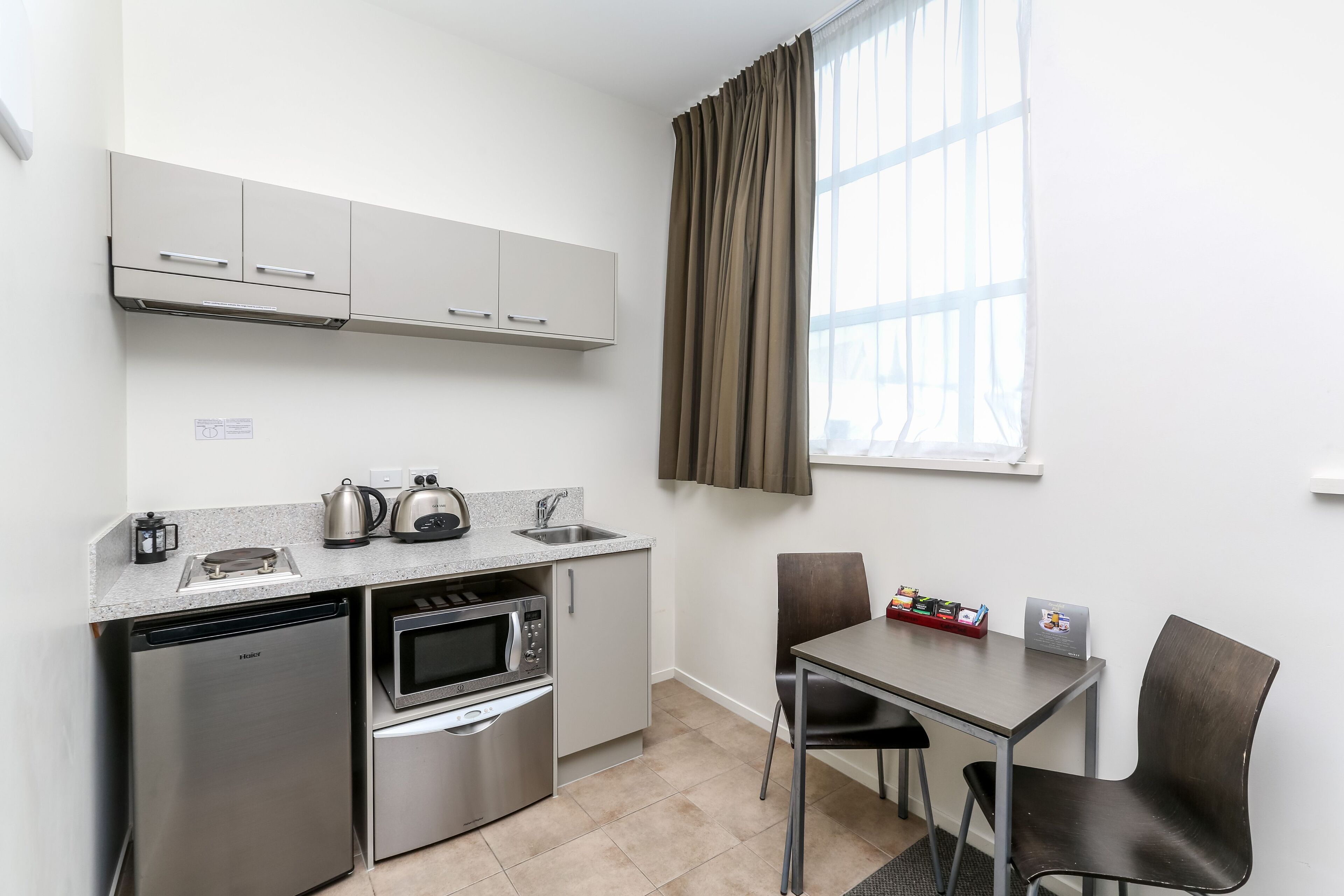 A compact kitchenette with a dishwasher, microwave, kettle, and sink. There is a small table with chairs and a window with curtains.