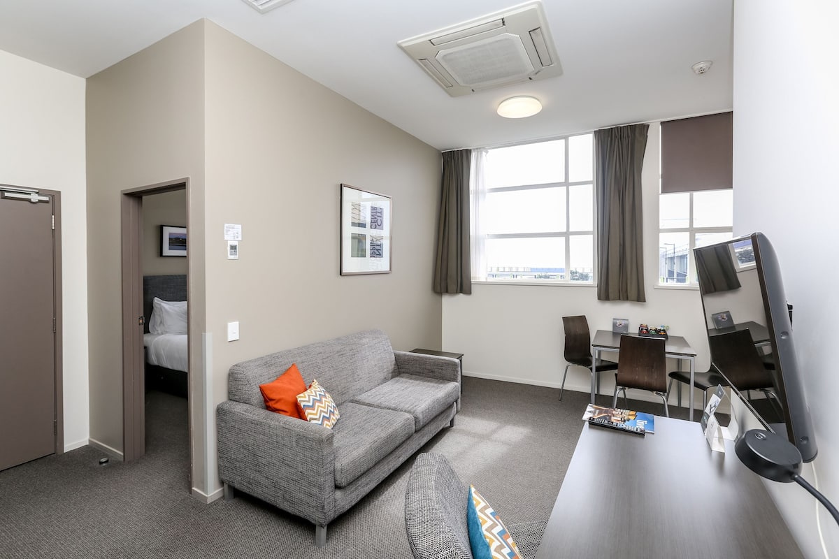 A modern hotel room with a grey sofa, a desk with a computer, a chair, and a large window with curtains.