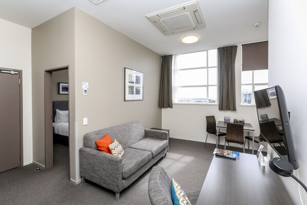 Habitación de hotel moderna con un sofá gris, un escritorio con computadora, una silla y un ventanal con cortinas.