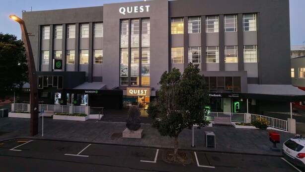 A modern building with a prominent 'QUEST' sign, a parking area, and a tree in front.