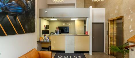 Zona de recepción de un hotel con un cartel 'QUEST', una recepcionista, un mostrador de recepción, un sofá con un tapete 'QUEST' y un ascensor.