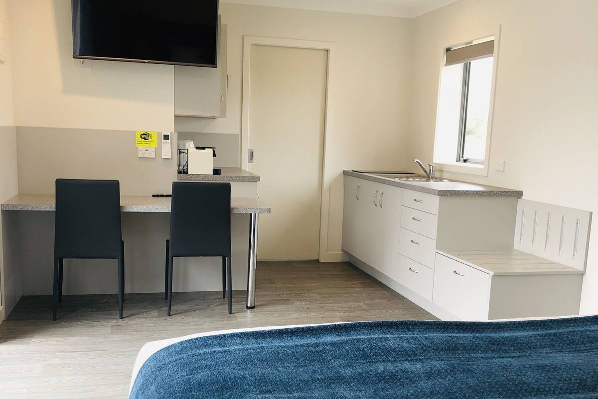 A modern hotel room with a bed, a desk, two chairs, a TV, a window, and a kitchenette.