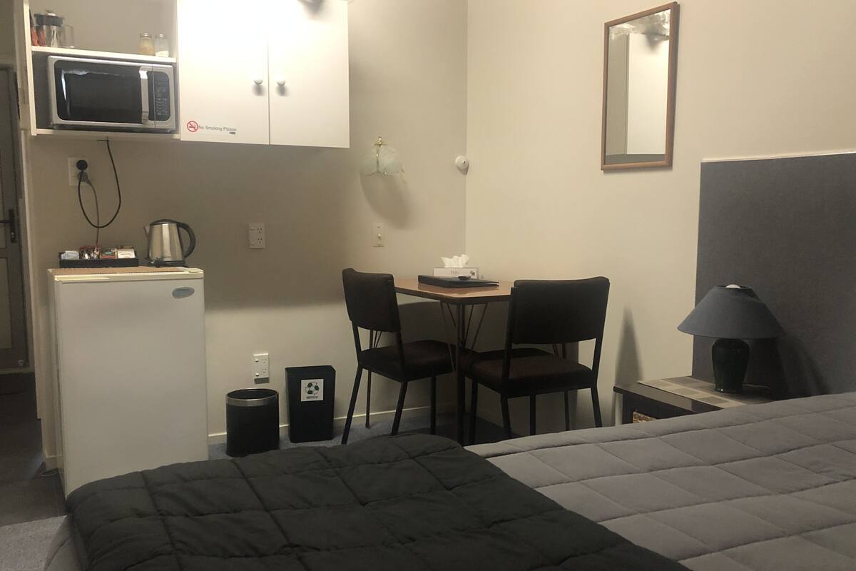 A small hotel room with a bed, a desk with two chairs, a microwave, a refrigerator, a trash bin, and a bedside lamp.