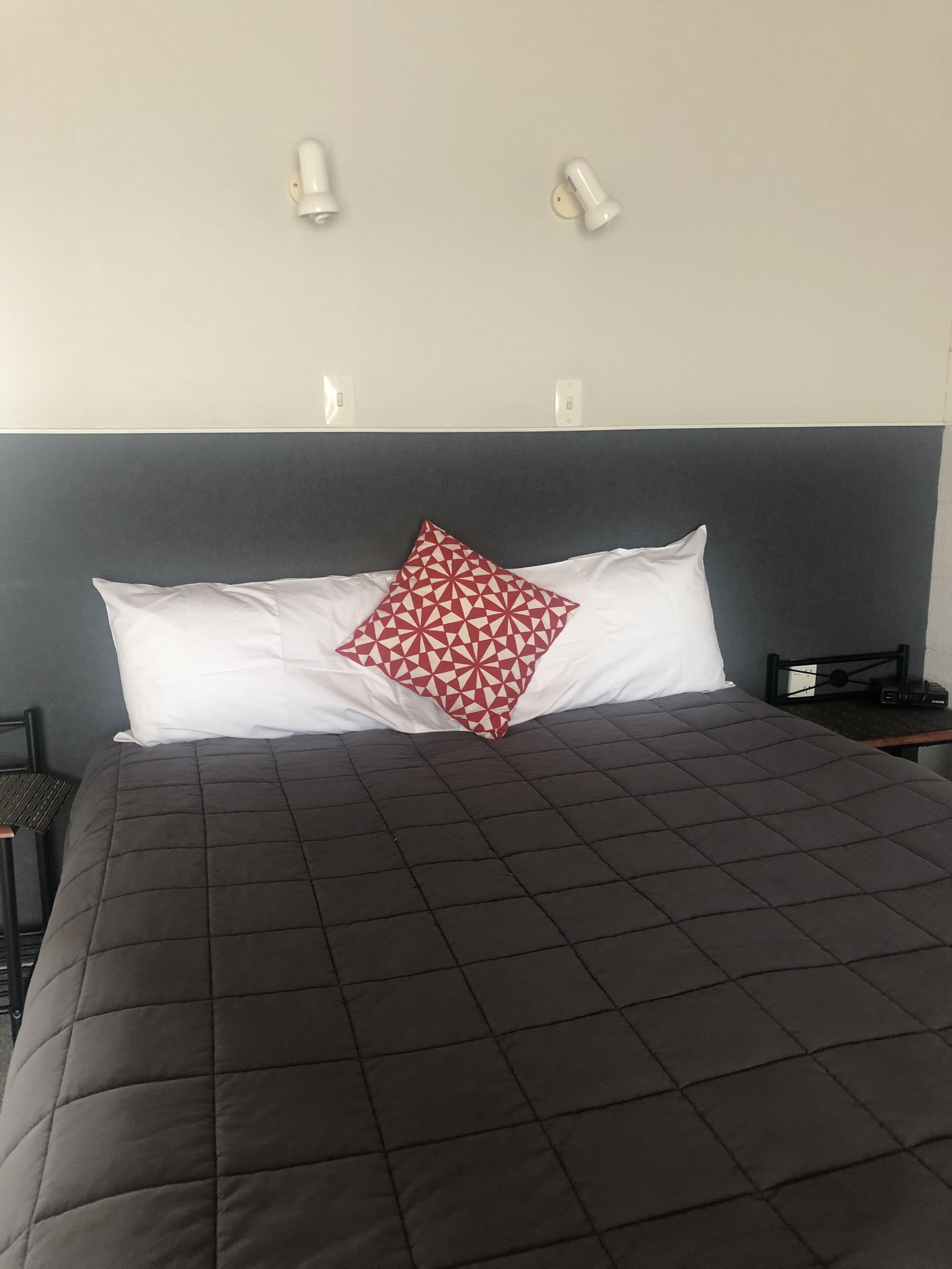 Un lit bien fait, recouvert d’une couverture sombre, avec un oreiller blanc et un oreiller rouge à motifs géométriques. Deux lampes murales sont fixées au-dessus du lit.