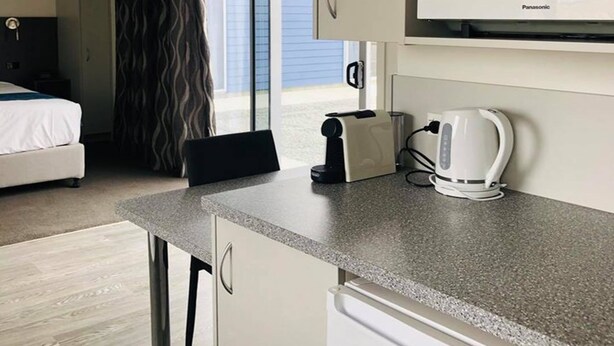 A compact hotel room with a kitchenette, a bed, and a view through a window. - Thornton Lodge Motel (Waipukurau)