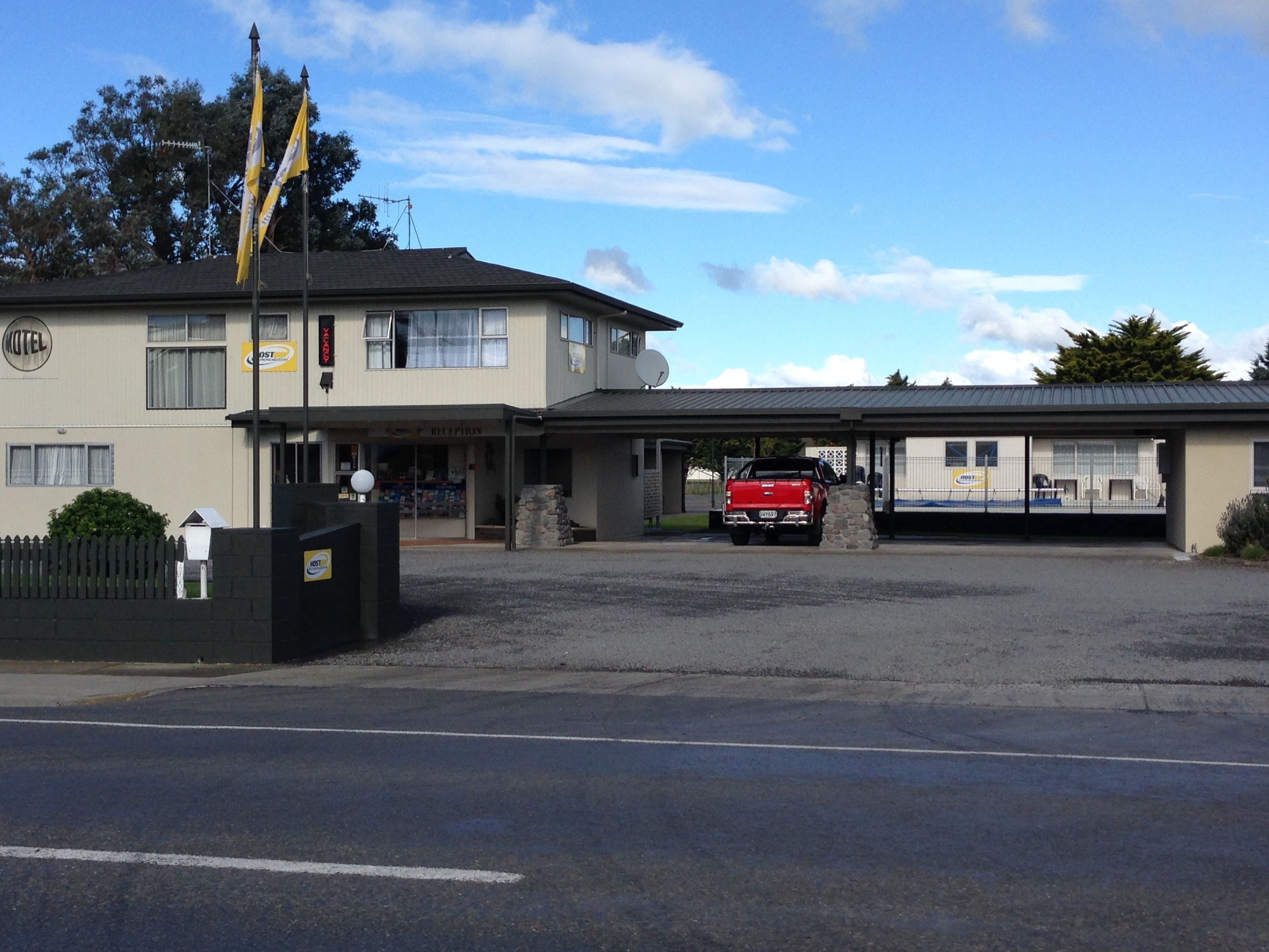 Un motel à deux étages avec un comptoir d’accueil, un camion rouge garé devant et un espace de stationnement clôturé.