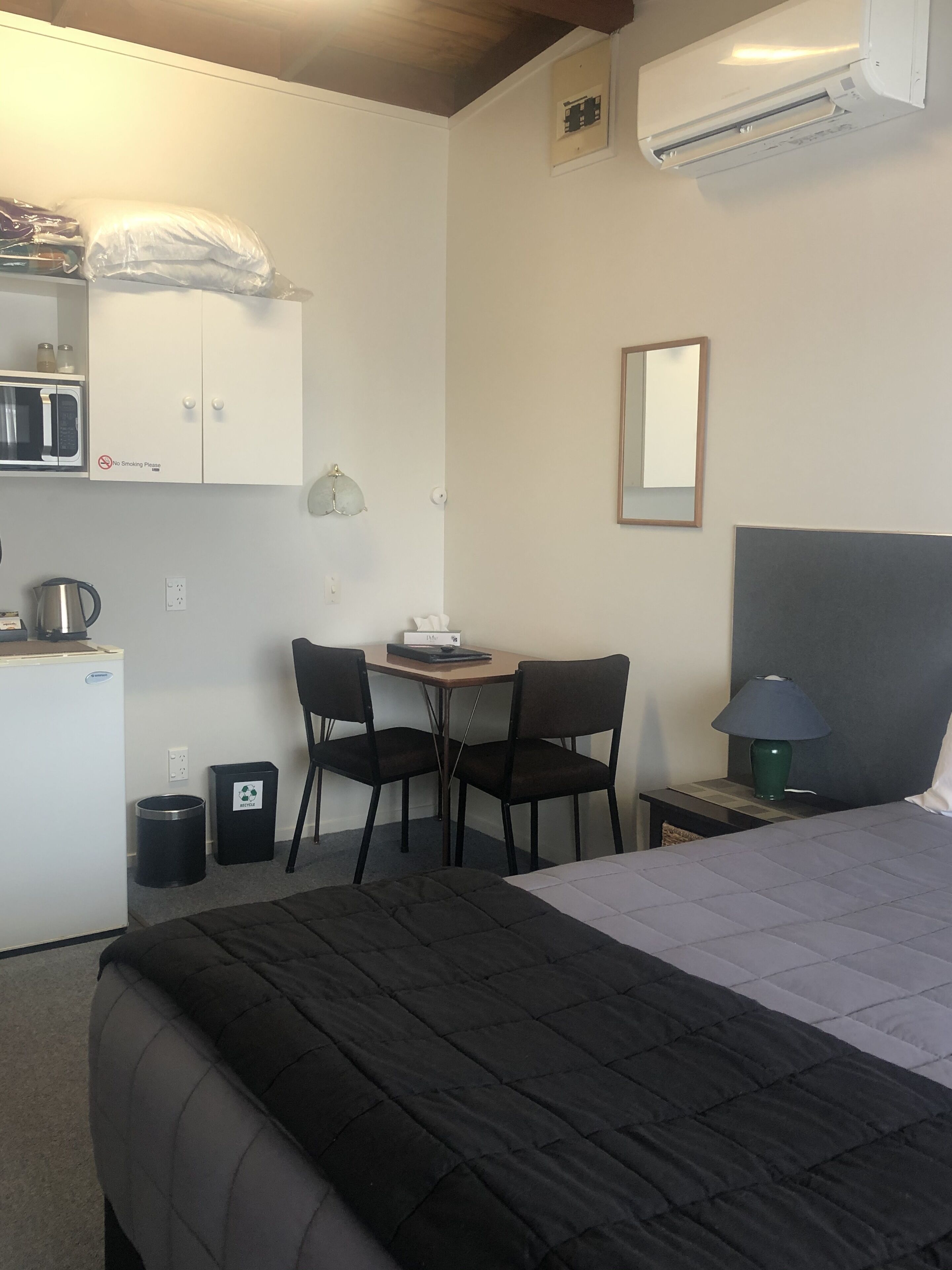 Quarto de hotel compacto com cama, pequena escrivaninha com cadeiras, micro-ondas, lixeira e ar-condicionado fixado na parede.