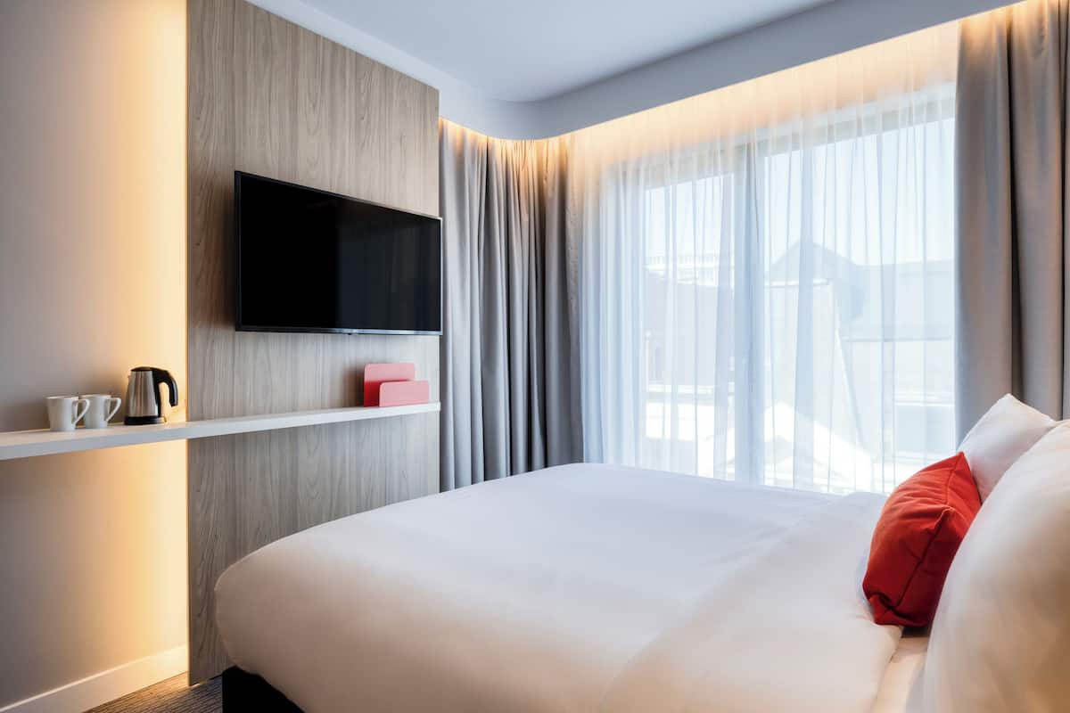 A hotel room with a bed, a mounted TV, a shelf with a kettle and cups, and a window with curtains.