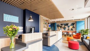 A hotel reception area with a wooden reception desk, a blue wall with a Holiday Inn Express sign, a bar area with shelves of bottles, and a seating area with orange chairs and a red ottoman. - Holiday Inn Express Antwerp City Centre by IHG (Antwerp)