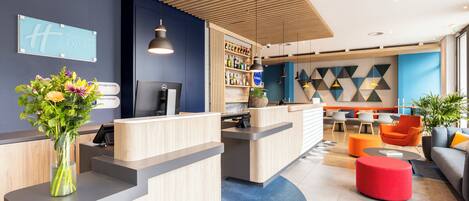 A hotel reception area with a wooden reception desk, a blue wall with a Holiday Inn Express sign, a bar area with shelves of bottles, and a seating area with orange chairs and a red ottoman.